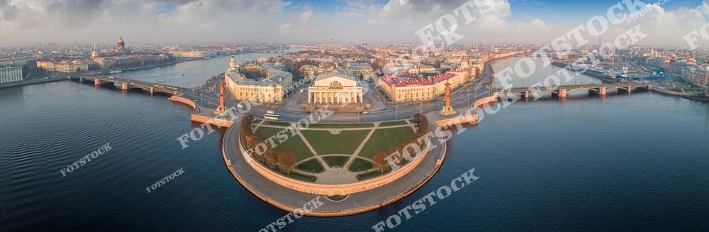 На фотографии изображена Стрелка Васильевского острова, один из символов Санкт-Петербурга. Ростральные колонны и здание Биржи гармонично сочетаются с набережными рек Невы и Малой Невы. С высоты открывается впечатляющий вид на исторический центр города, включая Исаакиевский собор и другие архитектурные памятники. Легкие облака и утреннее освещение создают атмосферу спокойствия и величия.